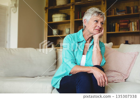 Senior woman sitting on sofa wearing turquoise shirt, gazing to right at home with wooden bookshelf 135307338