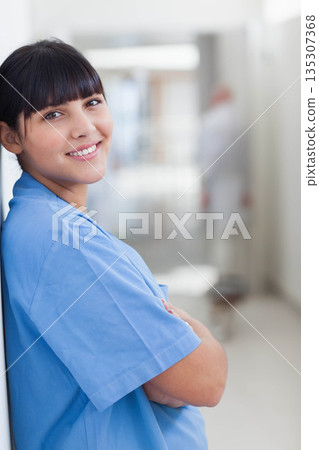 Female nurse leaning against hospital corridor wall, wearing blue scrubs and smiling, copy space 135307368