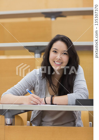 Asian woman smiling while holding blue pen over paper at lecture hall desk with binder Asian woman smiling while holding blue pen over paper at lecture hall desk with binder 135307370