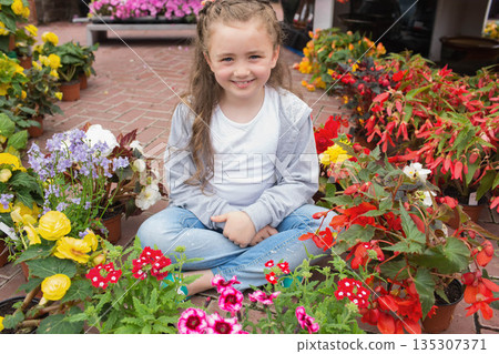 Elementary school girl sitting cross-legged among colorful potted flowers at nursery walkway 135307371