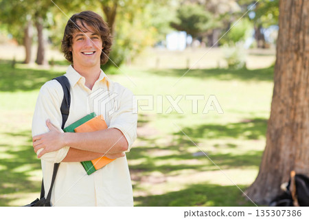 Male student standing on campus lawn holding green orange textbooks carrying backpack, copy space Male student standing on campus lawn holding green orange textbooks carrying backpack, copy space 135307386