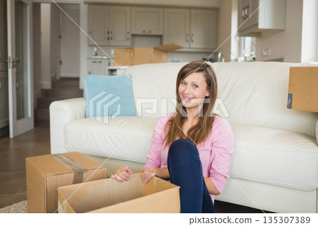 Woman sitting on floor packing moving boxes beside sofa in living room near kitchen cabinets 135307389