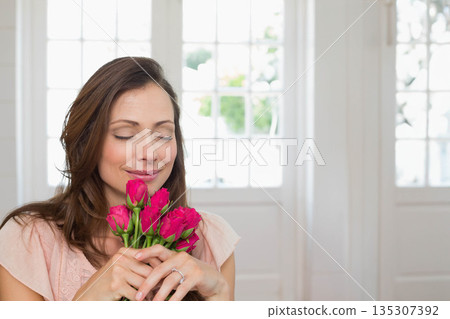 Mid adult woman standing at doorway holding pink roses close to face, copy space Mid adult woman standing at doorway holding pink roses close to face, copy space 135307392