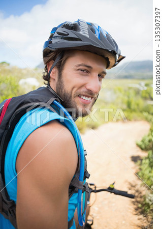 Male mountain biker standing on dirt trail glancing back wearing helmet and hydration pack 135307397