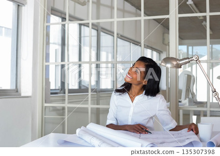 African American woman reviewing blueprints on desk in modern office under lamp with pen 135307398
