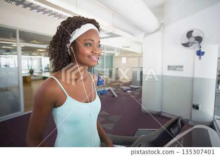 African American woman walking on treadmill at gym wearing tank top and earbuds, copy space 135307401
