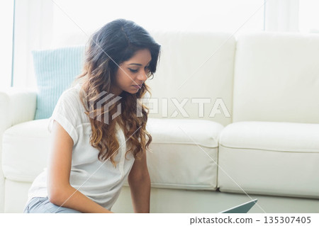 Asian woman kneeling wearing white blouse working on laptop in living room with sofa cushion 135307405