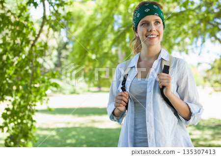 Woman standing beneath trees in sunlit park, wearing bandana and carrying backpack, copy space 135307413