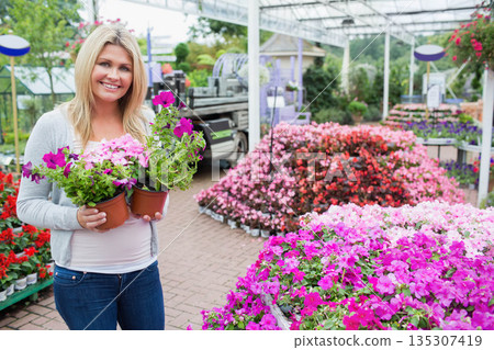 Woman holding two terracotta pots with vibrant petunias on brick path inside greenhouse, copy space Woman holding two terracotta pots with vibrant petunias on brick path inside greenhouse, copy space 135307419