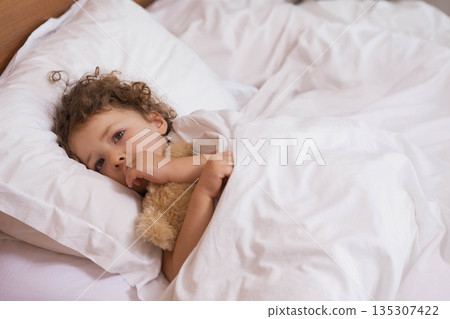 Girl with curly hair wearing pajamas hugging teddy bear lying on bed in bedroom, copy space Girl with curly hair wearing pajamas hugging teddy bear lying on bed in bedroom, copy space 135307422