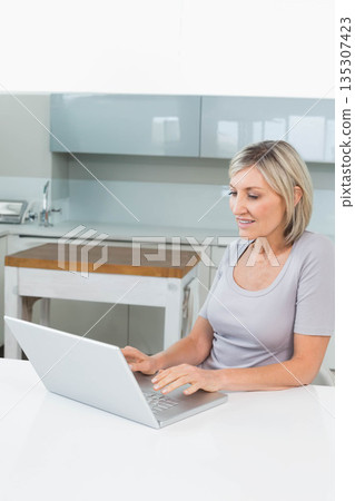 Woman typing on laptop at home kitchen table with chopping block, cabinets and faucet, copy space Woman typing on laptop at home kitchen table with chopping block, cabinets and faucet, copy space 135307423