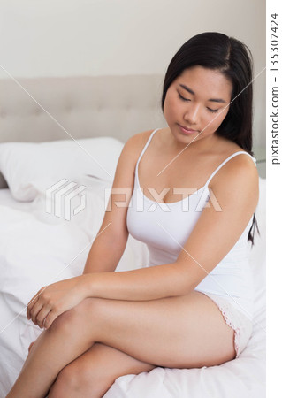 Asian woman sitting on bed wearing white camisole and pink lace shorts in bedroom, copy space 135307424