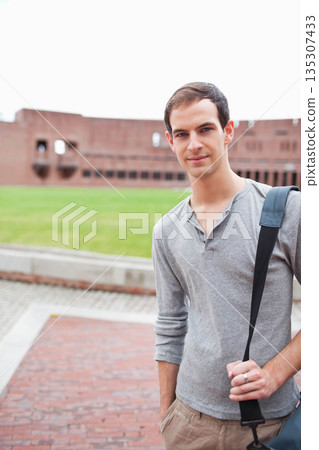 Male student standing in courtyard beside red brick building holding bag strap, copy space Male student standing in courtyard beside red brick building holding bag strap, copy space 135307433