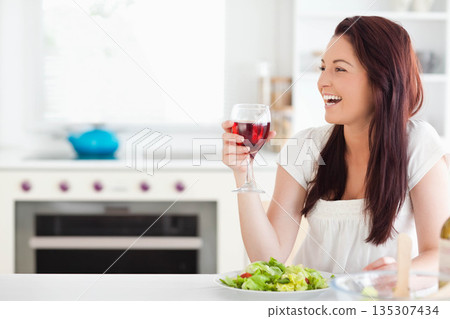 Woman holding wine glass while smiling at modern kitchen counter with salad plate, copy space Woman holding wine glass while smiling at modern kitchen counter with salad plate, copy space 135307434