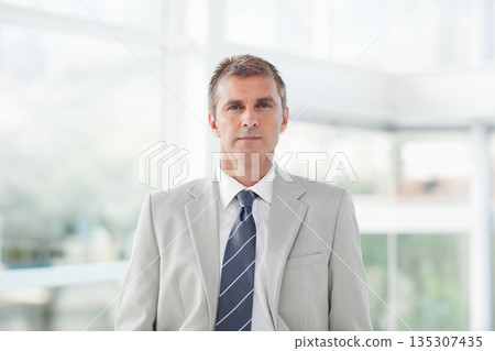 Middle-aged man standing wearing gray suit and striped tie in bright office lobby with glass walls 135307435