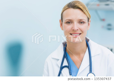 Smiling female doctor wearing white coat over blue scrubs with stethoscope in clinic, copy space Smiling female doctor wearing white coat over blue scrubs with stethoscope in clinic, copy space 135307437
