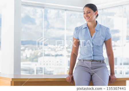 Woman sitting on wooden desk in office by floor-to-ceiling windows showing cityscape, copy space 135307441