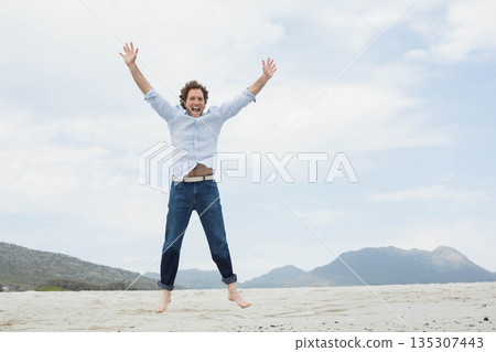 Mid adult man jumping on sandy shoreline wearing light blue shirt and dark jeans barefoot Mid adult man jumping on sandy shoreline wearing light blue shirt and dark jeans barefoot 135307443