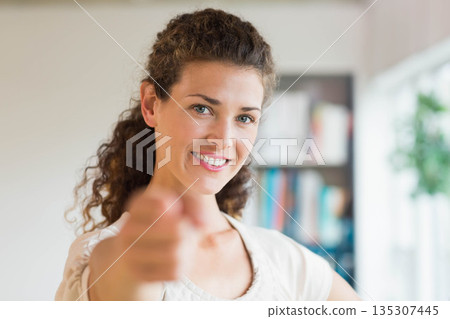 Woman standing and pointing forward in home study by bookshelf with books, window and potted plant Woman standing and pointing forward in home study by bookshelf with books, window and potted plant 135307445
