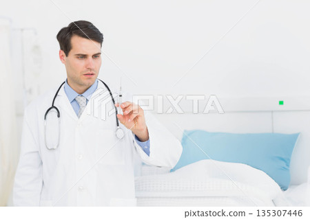Male doctor preparing syringe and wearing stethoscope beside hospital bed in exam room, copy space 135307446