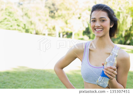 Asian woman holding clear water bottle standing on sunny park field in workout wear, copy space 135307451