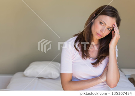 Woman sitting on bed edge touching temple near white pillow and woven basket, copy space Woman sitting on bed edge touching temple near white pillow and woven basket, copy space 135307454