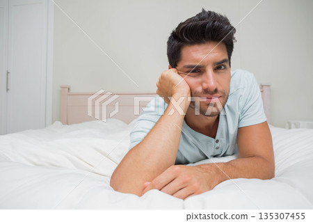 Male lying on bed with white comforter, head resting on hand, gazing toward camera in bedroom Male lying on bed with white comforter, head resting on hand, gazing toward camera in bedroom 135307455