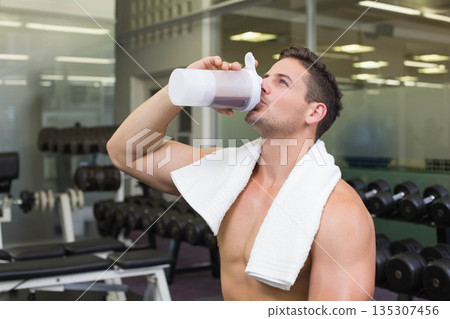 Shirtless male with white towel standing in dumbbell area of gym raising translucent shaker bottle 135307456