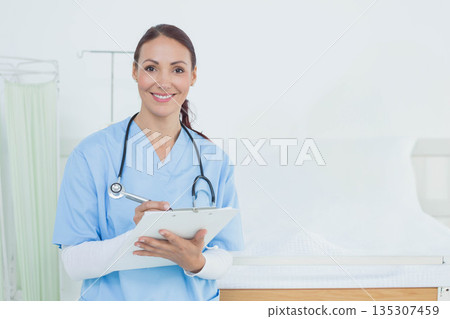 Middle-age woman wearing scrubs standing in clinic exam room holding clipboard and stethoscope Middle-age woman wearing scrubs standing in clinic exam room holding clipboard and stethoscope 135307459
