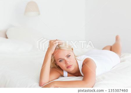 Woman wearing top lying on bed propping forearms on pillow beside lamp gazing sideways in bedroom 135307460