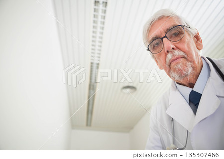 Senior doctor leaning toward viewer in exam room wearing lab coat with stethoscope, copy space 135307466