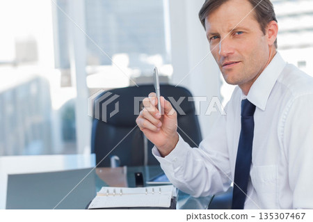Businessman holding pen at office desk with binder and tablet and looking toward camera, copy space Businessman holding pen at office desk with binder and tablet and looking toward camera, copy space 135307467