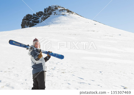 Woman carrying pair of alpine skis on shoulder wearing ski jacket on snow-covered slope, copy space Woman carrying pair of alpine skis on shoulder wearing ski jacket on snow-covered slope, copy space 135307469