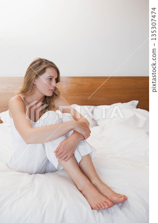 Woman wearing pajamas sitting on bedroom bed with headboard and pillows hugging knees, copy space 135307474