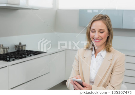 Businesswoman smiling while holding smartphone checking messages near stove in kitchen, copy space 135307475