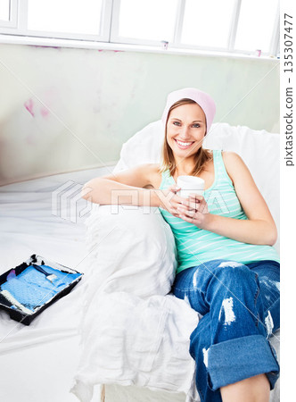 Woman sitting on sofa holding white mug during living room renovation with paint tray and roller 135307477
