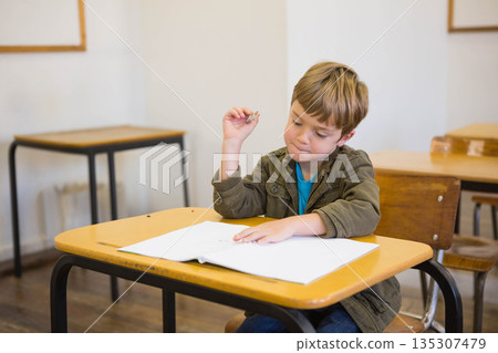 School-age boy sitting at desk in classroom, focusing on workbook holding pencil wearing jacket 135307479