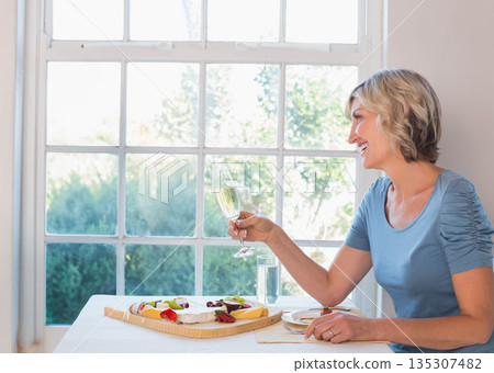 Mature woman sitting at dining nook holding sparkling beverage glass near serving board, copy space Mature woman sitting at dining nook holding sparkling beverage glass near serving board, copy space 135307482