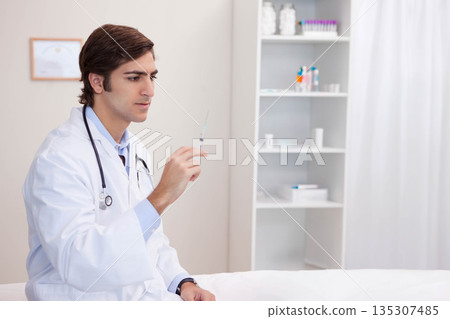 Male doctor inspecting syringe while wearing stethoscope on exam table in clinic room, copy space Male doctor inspecting syringe while wearing stethoscope on exam table in clinic room, copy space 135307485