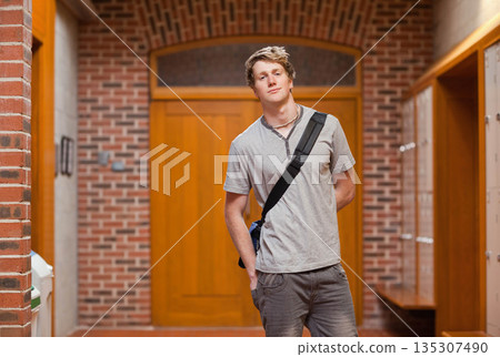 Male student standing in corridor wearing canvas shoulder bag near metal lockers, double doors 135307490