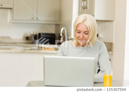 Woman browsing on laptop in modern kitchen while sipping orange juice beside sink 135307504