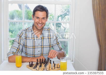 Middle-aged man sitting at dining table by window moving chess pieces near glasses of orange juice Middle-aged man sitting at dining table by window moving chess pieces near glasses of orange juice 135307533