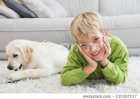 Blond boy lying on white shag rug at home smiling while puppy chewing bone near sofa Blond boy lying on white shag rug at home smiling while puppy chewing bone near sofa 135307557