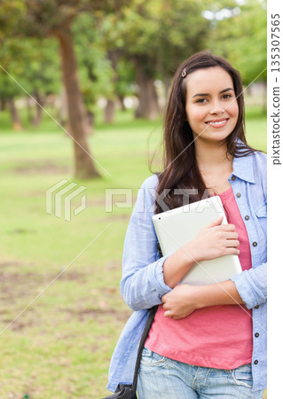 Female student standing in grassy park holding tablet computer and wearing backpack, copy space 135307565