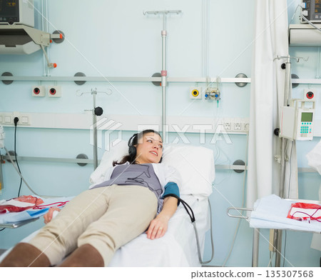 Reclining on hospital bed wearing headphones and blood pressure cuff near IV pole with tubing Reclining on hospital bed wearing headphones and blood pressure cuff near IV pole with tubing 135307568