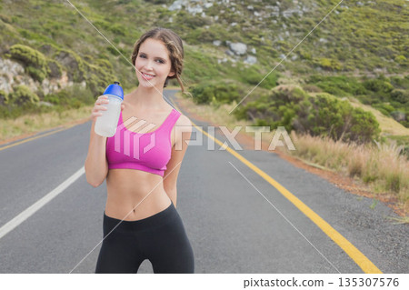 Female standing on winding highway wearing pink sports bra with black leggings holding water bottle 135307576