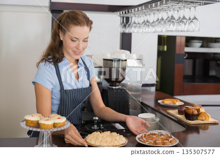 Mid adult barista arranging pastries next to coffee grinder at cafe counter wearing pinstripe apron Mid adult barista arranging pastries next to coffee grinder at cafe counter wearing pinstripe apron 135307577