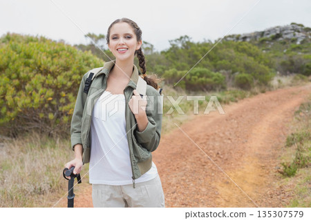 Woman hiker holding trekking pole while adjusting backpack on winding dirt trail, copy space 135307579