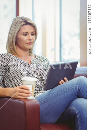 Woman sitting in leather armchair at home beside windows holding digital tablet and sipping coffee 135307582