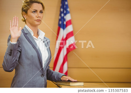 Woman wearing suit raising hand, placing hand on book at witness stand with flag, copy space Woman wearing suit raising hand, placing hand on book at witness stand with flag, copy space 135307583